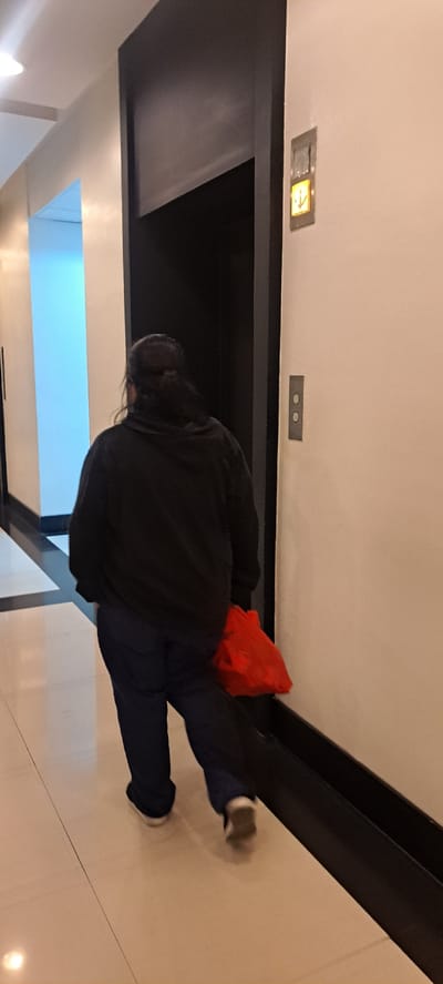 A person with dark hair pulled back in a ponytail walks away from the camera down a brightly lit hallway towards an open elevator. They are wearing a black hooded sweatshirt and dark blue pants. They are carrying a red shopping bag in their right hand, which is dragging slightly on the floor. The hallway has cream-colored walls with black trim along the floor. An elevator door, which appears to be dark, is open to the left of the person. An elevator call button with a lit downward arrow is visible on the wall to the right of the elevator entrance. The floor is tiled with a light beige color. This scene is located in the city of Mandaluyong, Philippines, likely within a building.