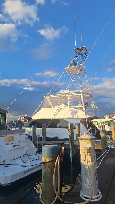 This image depicts a marina scene, likely in Nantucket, United States of America, given the context of the provided crops. The foreground features a wooden dock with several pilings, one of which is wrapped with a thick rope and a coiled hose. A utility pedestal, possibly for electrical and water hookups, is positioned on the dock, with cables connected.
In the mid-ground, two white boats are docked. The boat on the left appears to be a smaller vessel with "EAST GREENWICH, RI" visible on its stern. To its right, a larger, more prominent white yacht is moored. This yacht has a tall, multi-tiered structure with numerous support poles and rigging, indicative of a fishing or cruising vessel. The yacht's cabin is enclosed with clear plastic sheeting.
The background shows other boats and buildings, including structures with peaked roofs and a steeple, suggesting a waterfront town. The sky above is a vibrant blue, dotted with scattered white clouds, and illuminated by warm sunlight, indicating it is either early morning or late afternoon. The overall impression is a peaceful maritime setting.