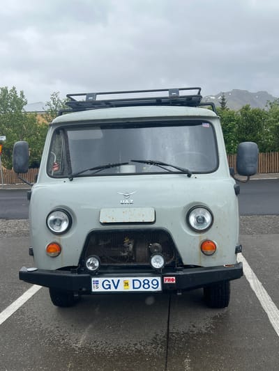 The image shows a light gray UAZ van parked in a paved parking lot in the town of Grundarfjörður, Iceland. The van has a roof rack, a front bumper with a black grille and two circular headlights on each side. The windshield is slightly wet. The van has an Icelandic license plate that reads "GV D89" with a small sticker below it that reads "PHØ." The van is facing the camera. The background shows buildings to the left and a mountain range to the right. The ground is wet from rain. The sky is overcast. The coordinates 64.926, -23.257 correspond to a location in Grundarfjörður, Iceland. The van is positioned in a parking lot near a street. The van's driver's side door is slightly open, the door handle is visible. The van is parked next to a white line. The image was taken on a cloudy day. There is no one present in the image.