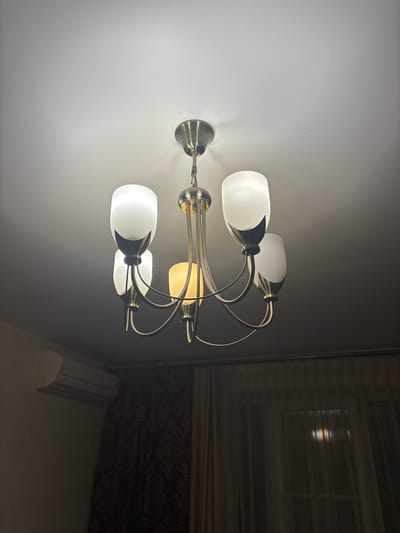A brass chandelier with five frosted glass globes hangs from a white ceiling. Four of the globes are illuminated with white light, while one is lit with a warm yellow glow. The chandelier is suspended by a chain and has a decorative central element. In the background, to the left, a white air conditioner unit is mounted on a tan wall. To the right, dark patterned curtains hang over a window, with a faint reflection of light visible on the glass. This scene is likely indoors, in a residential setting. The city of Otradnoye, Russia is not visible in this image.