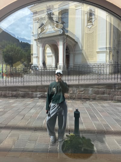 The image is a reflection in a curved window, showing a person standing in front of a building and a stone-paved area. The person is wearing a dark green jacket, grey pants, and a white cap. They are looking towards the camera. In the reflection, the building appears to be made of light-colored stone with arched entrances and ornate architectural details. A black metal fence is visible in front of the building. The stone-paved area is made up of rectangular bricks. The time of day appears to be daytime, with bright lighting and shadows suggesting a sunny day. The reflection also shows a glimpse of a mountainous landscape with green trees and a cloudy sky, indicating a natural setting. The overall mood is casual and perhaps a bit artistic due to the reflective surface. There are no discernible words or text in the visible portion of the image.