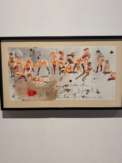 This is a framed artwork depicting a series of figures in a stylized, illustrative manner. The subjects appear to be human figures, predominantly female, depicted in various poses, some of which are acrobatic or contorted. They are rendered with bold outlines and colored with watercolors, giving them a somewhat abstract yet figurative quality. Some figures are nude or semi-nude, wearing lingerie or suggestive clothing. The background of the artwork is a textured paper with scattered ink splatters and watercolor washes, creating a somewhat chaotic or abstract environment.
To the right of the figures, there is handwritten text in a script style, which is difficult to decipher fully. Some words or phrases appear to be present, but their meaning or content cannot be determined from the image alone. The overall impression is that of a dynamic composition, possibly exploring themes of the human form, movement, or sensuality. The artwork is framed with a black border and a cream-colored mat. The setting appears to be an indoor space, possibly a gallery or home, where the artwork is displayed on a textured white wall. The lighting suggests an indoor environment, possibly during the day or with artificial lighting. There are no specific location cues within the artwork itself, other than the stated context of La Cuesta, Spain.