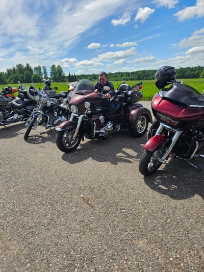The image shows a line of motorcycles parked on a paved road. A man is sitting on one of the motorcycles, looking at the camera. The background consists of a green field and a blue sky with clouds. The location is possibly near Borodinskogo Muzeya, Russia, given the presence of motorcycles suggesting a gathering or event.