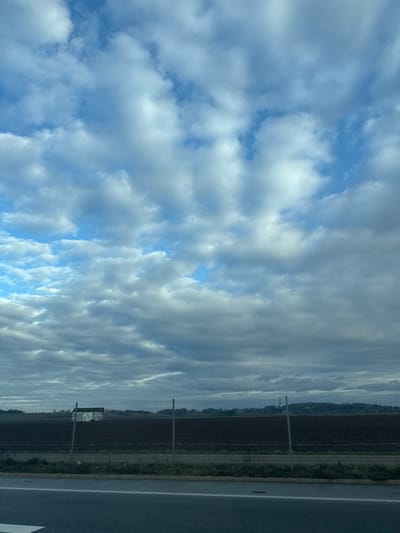 The image depicts an open landscape in Wolfisheim, France, under a cloudy sky. The upper two-thirds of the frame consist of a sky filled with various shades of gray and white stratocumulus clouds, with several patches of clear blue sky visible. The lower third of the image is divided horizontally into three main layers: a dark gray asphalt surface with a white painted line at the very bottom, followed by a low-profile wire fence with thin metal posts spanning the width of the frame, and then a wide expanse of dark brown, tilled earth. In the far background, beyond the cultivated field, a faint treeline is discernible, with a small, white-colored building featuring a dark roof visible on the horizon line towards the left. No human figures or active subjects are present in the scene.