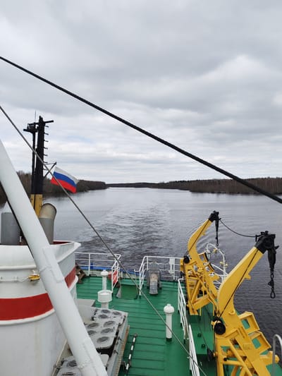 The image captures a view from the stern of a ship traveling along a wide river or canal, likely in Uzhla, Russia. The sky is overcast with a uniform layer of gray clouds, indicating a cloudy day. Both banks of the waterway are densely forested with bare deciduous trees, suggesting the season is late autumn or early spring.
In the foreground, elements of the ship's structure are visible. The deck is painted a practical green. On the left, a portion of the ship's white hull, accented with a red horizontal stripe, and a thick white mast or support pole are prominent. Further left, a darker mast-like structure displays a Russian flag, horizontally striped in white, blue, and red, fluttering in the breeze.
The right side of the deck is dominated by two robust, bright yellow crane-like structures, possibly davits, equipped with chains and hooks, designed for launching or retrieving objects. Visible text on one of these yellow components reads "ИСПОЛ.022-L" and "ТПРЭ.02.26", likely identifying markings or specifications. Other deck features include white railings, various metallic hatches with circular closures, and a white ventilation pipe.
The ship's movement is evident from the distinct wake trailing behind it in the dark water. Several thick, black ropes or cables belonging to the ship's rigging stretch diagonally across the upper portion of the frame. The river extends into the distance, bordered by the unbroken treeline on either side, conveying a sense of progression through a natural, inland waterway.