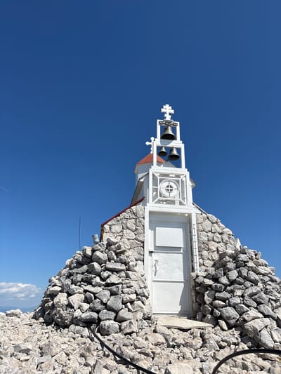 A low-angle, slightly rotated, outdoor shot depicts a small, white building constructed of stone and painted metal components, set against a clear, bright blue sky. The building is situated atop a rock formation. At the far left of the building is a white, metal structure supporting three small bells and topped with a Christian cross. To the right of the bells is a white, rectangular structure featuring a circular design. Adjacent to this is a square, metal panel with a diamond-patterned surface. To the right of the metal panel are two white, metal doors set into the stone wall of the building. A small portion of a red roof is visible on the side of the building. An electrical cable runs along the top right of the stone structure. The location is deduced to be Mali Mikulići, Montenegro, based on the architectural style and landscape.