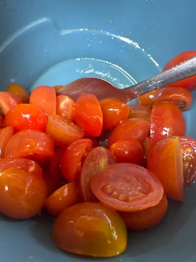 The image depicts a close-up, overhead view of numerous cherry tomatoes, predominantly halved lengthwise, contained within a light blue bowl. The tomatoes display varying shades of red and reddish-orange, with some pieces exhibiting a subtle yellowish-green hue, and possess a wet, glossy surface texture. A silver-colored spoon is partially visible, resting among the tomatoes inside the bowl. The scene is located in Invergowrie, United Kingdom.