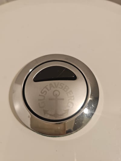 This media file is a close-up, eye-level shot of a toilet flush button. The button is round and made of chrome-plated metal, with a smaller black rectangular insert on the left side. The button has text engraved on its surface. The visible text reads "GUSTAVSBERG," with a graphic symbol resembling a stylized "G" and a plus sign or cross in the center. The button is mounted on a white ceramic toilet tank. The lighting suggests an indoor setting, possibly a bathroom, with some reflections visible on the chrome surface. The image appears to have been taken in Mäntsälä, Finland. There are no people or discernible activities taking place, focusing solely on the object itself.