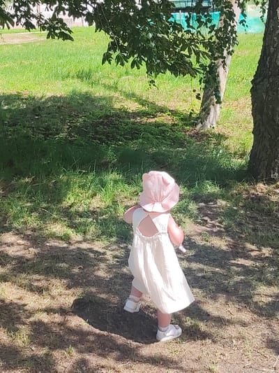 A young child wearing a light-colored dress and a pink sun hat with ear details stands on a patch of dry grass in a park. The child is facing away from the camera, looking towards a grassy area with trees. Sunlight casts shadows on the ground. The background includes a tree trunk, green grass, and foliage from a larger tree in the foreground. The scene appears to be outdoors on a sunny day.