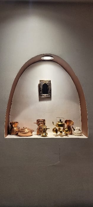 An alcove in a wall displays decorative items. A small, ornate black-centered frame hangs to the left. To the right are multiple metallic, possibly brass or copper, vessels. These seem to be arranged vertically, varying in size and shape, from small cups and pots to larger, lidded containers. The alcove's interior is bright, while the surrounding wall is darker. A light fixture is visible within the alcove's curved top.