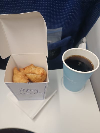 A close-up shot shows a meal served on a tray table in what appears to be an airplane or train. In the foreground, a white cardboard box with a blue border contains two golden-brown samosas. The box has "Flying Samosa" printed on it in blue lettering. To the right of the box is a light blue paper cup filled with a dark beverage, possibly coffee or cola. The cup has text that is partially obscured but appears to say "Hydrated." The background reveals a blue, textured seat, typical of an airplane or train, with a mesh pocket visible on the side. The tray table is white. The lighting is artificial and casts soft shadows. There are no people visible in the image. The scene is presented in a way that suggests a travel snack or meal. The location in the city of Agassaim, India is not indicated by any visual cues in the image.
