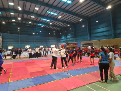 An indoor sports hall in Phagwara, India, hosts a kabaddi match. Red and blue mats delineate the playing field, marked with yellow and black lines. Several individuals, appearing to be participants and officials, are present on the court and surrounding areas. Some are dressed in athletic uniforms, while others wear casual attire. In the background, a large banner displays "KENDRIYA VIDYALAYA SANGATHAN 59TH NATIONAL SPORTS MEET." The hall features a high, corrugated metal roof with numerous lights and skylights. Spectators can be seen in the bleachers in the far background. The overall scene depicts an organized sporting event within a modern gymnasium.