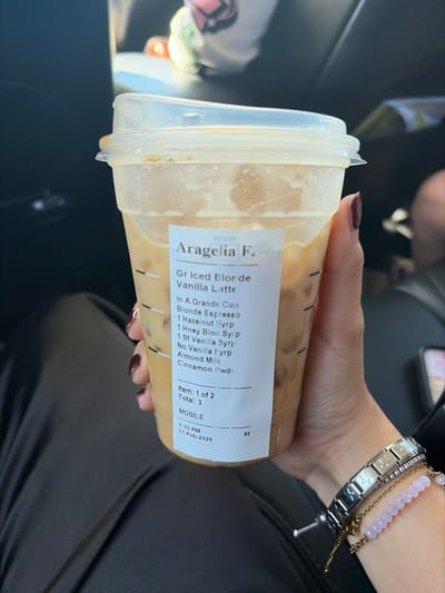 A hand holds a large, clear plastic cup containing a Starbucks iced latte. The label identifies it as a "Gr Iced Blor de Vanilla Latte" ordered for Aragelia F. and specifies a Blonde Espresso, Hazelnut, Honey Blend, and Sf Vanilla syrups, Almond Milk, and Cinnamon Powder. The order includes 3 items, with this drink being item 1 of 2. The mobile order was placed at 1:35 PM on February 7, 2026. The person's wrist sports a bracelet and a watch.
