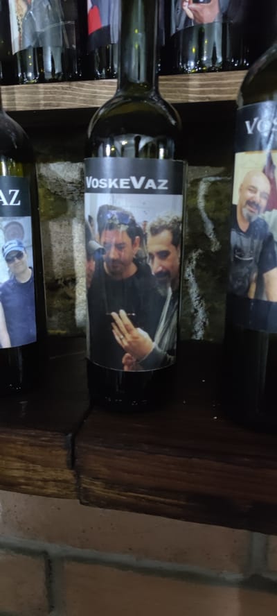 The image depicts a close-up view of three wine bottles arranged on a shelf, likely in Voskevaz, Armenia, given the prominent text "VoskeVaz" on the central bottle's label. The bottles are made of dark green glass and have labels featuring photographs of men.
The leftmost bottle has a partial label with a man wearing a blue cap and sunglasses. The central bottle has a label that reads "VoskeVaz" in white sans-serif font. Below this text, the label displays a photograph of two men. One man, in the foreground, is wearing a black shirt and appears to be looking at a phone held in his hand. The other man, to his right, has a beard and is also looking in the same direction. The third bottle, on the right, is partially visible and its label shows a close-up of a bald man with a beard, wearing a dark t-shirt.
The bottles are placed on a wooden shelf, which is positioned in front of a stone or brick wall. The lighting appears to be dim, creating shadows and highlighting the glossy surface of the bottles. The overall impression is that of a display or collection of wine bottles, possibly in a winery, shop, or a private collection, located in Voskevaz.