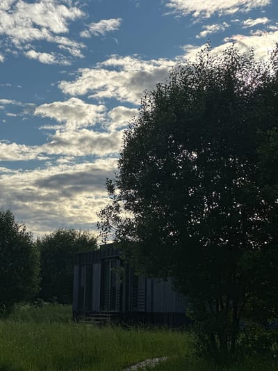 The image depicts a modern, dark-colored, rectangular structure with large windows, partially obscured by lush green trees and tall grass. The structure appears to be a dwelling or a small building. The sky above is a vibrant blue, dotted with scattered white and gray clouds, suggesting a partially overcast day. The overall setting is rural or semi-rural, with abundant vegetation. The location is identified as Znamenka, Russia. There are no people visible in the image.