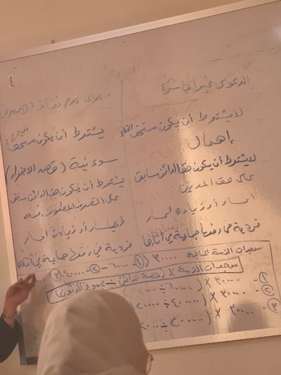 This image captures an indoor scene in Al-Kiswa, Syria, focused on a whiteboard during an instructional setting.

A large, white magnetic whiteboard with a silver frame is prominently displayed, covered with handwritten Arabic text and numerical entries in blue or black marker. Visible Arabic phrases include "الدعوى غير المباشرة" (The Indirect Lawsuit), "إهمال" (Negligence), "يشترط أن يكون" (It is required that it be), and "موجبات الذمة الحالية" (Current Liabilities). A rectangular box is drawn around a section of text and numbers on the lower-middle portion of the board, which also features circled numbers (e.g., ①, ②, ③, ④) alongside numerical values.

In the left foreground, the right arm and hand of an individual with light brown skin are visible, clad in a dark, long-sleeved garment. Their index finger points directly at the lower-left section of the whiteboard, specifically at the content within or near the drawn box.

In the immediate foreground, partially obscuring the lower-middle of the whiteboard, is the back of a second individual's head and shoulders. This person is wearing a light-colored hijab and glasses, facing towards the whiteboard, indicating observation or engagement with the material.

The scene is lit by indoor illumination, giving the image a slightly warm tone. The environment appears functional, consistent with a classroom or lecture hall.