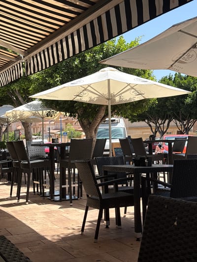 This media file depicts an outdoor seating area of a cafe or restaurant, likely in Campillo Del Moro, Spain. The scene is set during the daytime, with bright sunlight casting shadows across the paved patio. Several tables and chairs made of dark, woven material are neatly arranged, suggesting the establishment is either closed or preparing for service.

Dominating the foreground are large, cream-colored umbrellas providing shade. One umbrella prominently displays circular logos with text, though the specific brand is not clearly legible. Another umbrella in the background is partially obscured by trees. To the left, a striped awning, featuring black and white bands, extends over the seating area, adding a graphic element to the scene.

Behind the seating area, lush green trees provide a natural backdrop. In the distance, buildings with a light-colored facade and red-tiled roofs are visible, hinting at the architectural style of the locale. A white van and a red vehicle are also present in the background, suggesting a street or public area adjacent to the cafe.

The overall atmosphere is peaceful and tranquil, with no people visible. The neat arrangement of furniture and the lack of activity imply either an early morning, a siesta period, or that the establishment is not yet open. The clear sky and bright sunlight indicate fair weather. No explicit text beyond the logos on the umbrellas is clearly discernible.
