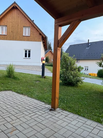 The image depicts an outdoor scene at dusk in Milná, Czechia. In the foreground, a paved patio made of rectangular grey stones is visible. To the right, a portion of a wooden post and roof structure, likely part of a porch or carport, is present. Beyond the patio and to the left, a lush green lawn with scattered white flowers extends.
In the mid-ground, a pathway of the same grey pavers leads away from the patio. Adjacent to the pathway stands a black lamppost with an illuminated yellow light. A cluster of tall green grass grows near the lamppost.
Further back, a white building with a wooden-paneled upper section and a dark tiled roof is situated. This building has several white-framed windows. To the right of this building, another white structure with a dark tiled roof and windows is visible, and beyond that, a partially visible wooden building.
The environment suggests a residential or rural setting. The sky is a soft blue, indicating twilight. There are no people or animals visible in the frame. The scene is static and peaceful.