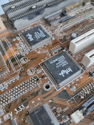 This is a close-up, eye-level, static shot of a dusty, vintage computer motherboard. The motherboard is predominantly brown with copper traces and features several integrated circuits, expansion slots, and other electronic components. One prominent black chip in the foreground has "Intel" branding and text that appears to include "PCIset" and a model number. Other visible components include white plastic expansion slot connectors, capacitors, and various small resistors and connectors. The overall condition of the motherboard suggests it is old and has been exposed to dust. The image does not depict any specific location in посёлок Областной сельскохозяйственной опытной станции, Russia, as it is a close-up of an electronic device.