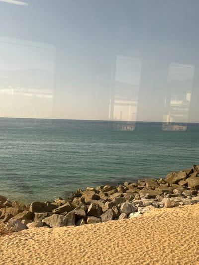 The image depicts a coastal scene in Cabrera de Mar, Spain, viewed through a window. The foreground features a sandy beach with footprints and scattered dried vegetation. Behind the beach, a rocky breakwater separates the sand from the sea. The sea itself is a vibrant turquoise color, with gentle waves. In the distance, the hazy horizon shows buildings and mountains, possibly part of the town or surrounding landscape. Reflections of a window frame and possibly interior elements are visible on the surface of the water and in the sky, suggesting the photograph was taken from inside a moving vehicle or building. The sky is a pale blue, with some atmospheric haze.