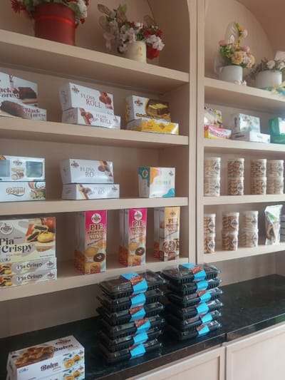 A bakery shelf displays an assortment of packaged goods. The top shelf features "Black Forest" cake, "Floss Roll," and other baked items. The middle shelf presents "Bogul" and "Floss Ro," with other pastries in boxes. The bottom shelf exhibits "Pia Crispy" and "Pie Brownies Susu." Stacks of brownies in black containers with blue labels are on the counter, alongside boxes of "Bolen" pastry. The shelves are light beige.