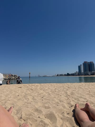 The image shows a beach on a clear day in Toseong-myeon, South Korea, with sand occupying the right half of the image. Buildings are visible along the coastline in the distance. The left half of the image is a clear blue sky. The sea is visible between the sand and the buildings. In the foreground, there are people near rocks, along with an umbrella. There are also legs and feet on the sand, indicating someone is relaxing on the beach.