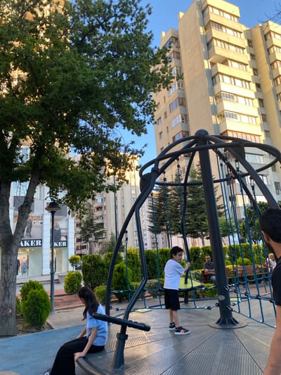 The image depicts a playground scene in front of a multi-story building, with lush green trees framing the view. The time of day appears to be late afternoon, as suggested by the warm, soft lighting and the shadows cast. The weather seems pleasant, with a clear blue sky visible above.
In the foreground, a circular metal playground structure with blue ropes is prominent. A young boy, dressed in a white t-shirt and black shorts, is actively engaged with the equipment, holding onto the ropes. A young girl, wearing a light blue t-shirt and black pants, is also interacting with the structure, sitting on one of its curved bars. In the background, another person is seated on a bench, observing the children.
The setting is urban, with a large, modern building dominating the background. The building features numerous windows and balconies, indicating a residential or commercial space. The trees in the foreground provide a natural contrast to the urban architecture.
The ground of the playground is a textured, metallic surface. Some signage is visible in the background, including "BAKER," suggesting a retail establishment. The overall mood is one of leisure and outdoor activity, with the children enjoying themselves on the playground.