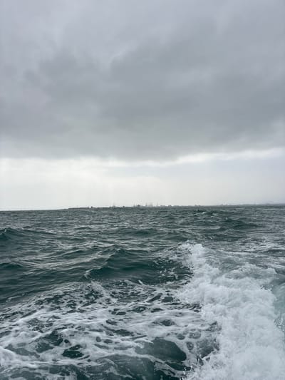The image depicts a choppy, grey ocean with a prominent wave breaking on the right side. The water is dark and textured, with white foam and spray from the wave. The horizon line is visible, separating the water from a hazy, overcast sky. In the far distance, along the horizon, a faint outline of a city skyline can be seen, with buildings appearing as small, dark shapes against the light sky. The overall mood is somber and somewhat dramatic, likely captured during a cloudy day with potentially rough seas. The location context of La Punta, Peru, suggests a coastal scene, possibly from a boat or pier, looking out towards the ocean and the city. There is no visible text in the image.