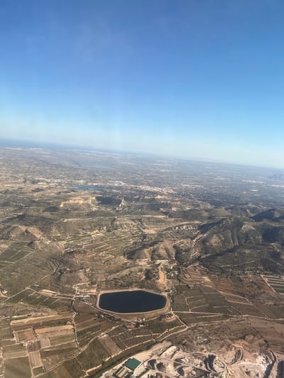 The image shows an aerial view of Aspe, Spain. The sky is clear blue on the left side. The land on the right side is mostly brown, with a mix of vegetation and agricultural fields. A large, dark, oval-shaped reservoir is visible. Near the upper right of the image there's a quarry/mining area. The landscape transitions from rural to urban areas as the view moves left.