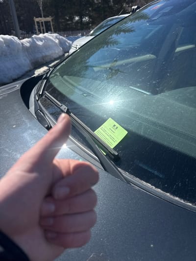 A forensic description of the scene in La Salle-les-Alpes, France:
The image captures an outdoor, daytime scene under clear skies, focusing on the front of a dark-colored vehicle, likely a car, in a winter or early spring setting. In the lower-left foreground, a human hand, specifically the left hand, is positioned giving a "thumbs-up" gesture. The vehicle's hood and a dust-covered windshield occupy the midground. A bright sun flare reflects prominently on the windshield's surface. A bright green, rectangular information slip or notice, bearing the word "INFORMATION" at its top and French text along with a French governmental coat of arms, is secured under the passenger-side windshield wiper. A Vehicle Identification Number (VIN), "YVZBW1A4AF2720118," is visible etched or printed on the lower section of the windshield near the dashboard. In the background, a significant, dirty snow pile runs along what appears to be a roadside or parking boundary. Partially visible beyond the snow is a portion of a second, lighter-colored vehicle. Sparse trees stand further in the distant background.