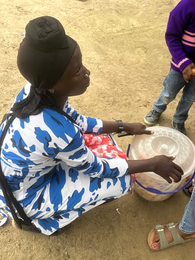 In Ganyingbo, Nigeria, a woman is kneeling on sandy ground, facing right in profile. She wears a black hijab styled into a bun and a white and blue camouflage-patterned long-sleeved dress. Her left hand rests on a clear plastic container with a blue rim, filled with food items. A child in a purple sweater with pink stripes and blue jeans stands to her right, holding an orange food item. Another person's legs and feet, wearing blue jeans and sandals, are visible in the foreground and to the right. The environment is dusty and appears to be an outdoor market or communal gathering space.
