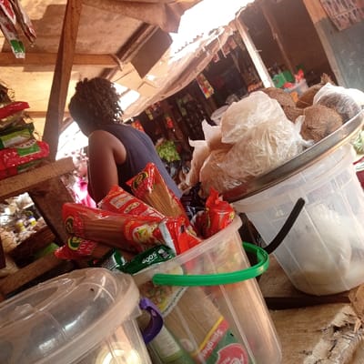 This image depicts a market stall scene in Obukpa, Nigeria. In the foreground, multiple transparent plastic buckets contain various food products. One large clear bucket, positioned centrally, holds numerous red-packaged pasta or spaghetti items, some labeled "Power Pasta" and "Spaghetti Pasta," alongside green-packaged goods; it features a green handle. To its right, another clear plastic bucket with a black handle supports a silver tray. This tray contains several whole brown coconuts and multiple small, clear plastic bags filled with a white, granular or stringy substance, likely food items. An additional clear plastic container with a purple handle is partially visible at the bottom left. These items rest on a rough wooden surface.
In the midground, a person with dark, curly hair, wearing a dark sleeveless top, is visible from the back, leaning forward toward the left. A second individual, wearing a red garment, is partially visible further in the background. The environment consists of a market structure with wooden beams and a corrugated metal roof, suggesting an outdoor or semi-sheltered market setting. Bright daylight illuminates the scene, casting shadows.