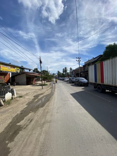This image captures a street scene in Lhoknga, Indonesia, on a sunny day with scattered clouds. The foreground features a paved road with a dirt shoulder on the left. Several vehicles are present, including a white truck with a colorful container, a dark-colored SUV, a few cars in a line, and a motorcycle carrying a rider in a yellow shirt moving away from the camera. Buildings line the right side of the road, and on the left, there are shops with signage, one of which reads "RACUN ETAR". Overhead, a complex network of power lines is visible against the blue sky. The environment appears to be a commercial or residential area with tropical vegetation, including palm trees, in the background. There are puddles of water on the dirt shoulder, suggesting recent rain or a moist environment.
