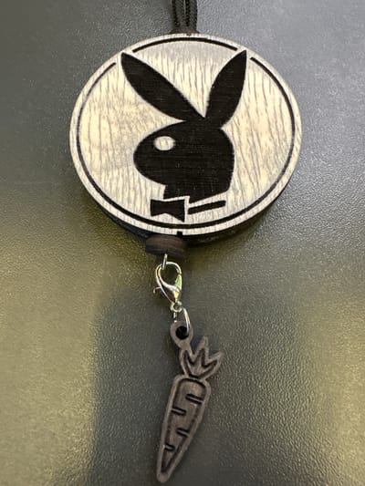 A close-up, top-down, angled shot shows a wooden keychain with the iconic Playboy bunny logo. The bunny is black, silhouetted against a light wood grain circle, and sports a black bow tie. Below the circular logo, a small, metallic clasp connects to a dangling charm shaped like a stylized carrot. The background is a dark, textured surface, possibly a table, with subtle lighting creating a slight sheen. The scene is static and offers no information about its location within Mikhailovka, Russia.