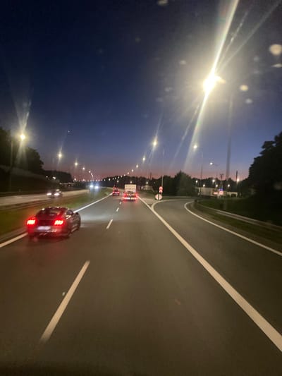 This media file depicts a nighttime drive on a highway in Zakrzów, Poland, during twilight. The scene is viewed from within a moving vehicle, looking out at the road ahead and the surrounding landscape.
The primary subjects are vehicles on the highway, including a black car in the foreground with its red taillights illuminated, and several other cars and trucks further down the road. The road itself is paved with prominent white lane markings. The setting is a highway with trees lining the left side and a faint silhouette of buildings or structures in the distance. Streetlights are visible along the highway, casting pools of light. The sky is a deep blue, transitioning to a lighter shade near the horizon, indicating twilight.
The activity taking place is a car journey on a highway at dusk. The headlights and taillights of the vehicles suggest movement and night driving conditions. The image is characterized by light flares from the headlights and streetlights, and some water droplets or smudges on the lens, adding a slightly ethereal quality to the scene.
Notable details include the time of day as dusk, indicated by the sky color and the use of headlights. The weather appears clear, though the presence of light flares might suggest some atmospheric moisture or a dirty lens. The location is identified as Zakrzów, Poland, though this is not explicitly visible in the image itself. There is no visible text in the image. The overall mood is one of quiet travel and observation during the transition from day to night.