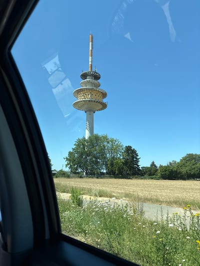 The image shows a telecommunications tower, likely the Feldbergturm, in Ober-Olm, Germany. The tower is tall and slender with multiple observation decks and a communication array at the top. It is situated in a rural area with a field of dry grass in the foreground, transitioning to a line of trees in the mid-ground. The sky is clear blue. The photograph appears to be taken from inside a vehicle, as the reflection of the car's interior and a portion of the car window frame are visible in the foreground. The scene is brightly lit by sunlight, suggesting it is daytime.