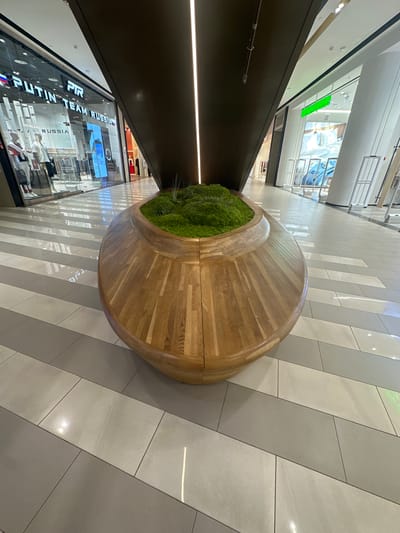 The image shows a high-angle view within a modern shopping mall or commercial space, likely in Родниковское сельское поселение, Ukraine. A large, circular wooden bench with a central patch of dense green plants serves as a focal point. The bench sits on a tiled floor with a diagonally striped pattern in shades of gray. Reflected light from the surrounding shops adds highlights to the tile surfaces.
In the background, there are storefronts featuring brightly lit interiors with visible displays of merchandise and branding, including the words "PUTIN TEAM RUSSIA." The lighting suggests a brightly illuminated indoor environment. The overall perspective gives a sense of the mall's layout and its integration of seating and green space within the shopping area. There are no people directly visible in the immediate foreground, but reflections in the glass storefronts suggest their presence.