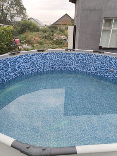 An outdoor above-ground swimming pool filled with clear blue water is the central focus of the image. The pool liner features a mosaic pattern of small blue and white tiles. The edge of the pool is visible, with a white top rail and dark grey supports. In the background, a grey concrete building with a white-framed window is on the right. To the left of the building and behind the pool, a yard is visible with overgrown grass, bushes, and trees. A pink and black cement mixer is present in the yard, along with scattered debris and some fencing. The sky is overcast. The scene appears to be in a rural or suburban setting in the city of Muhametjan Tüymebaev, Kazakhstan, likely a residential area given the presence of houses and the yard.