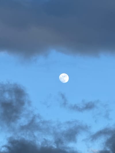 The image displays a full or nearly full moon, luminous white with discernible surface features, set against a vibrant blue daytime sky. The moon is positioned centrally within a clear patch of sky. Dark, heavy cloud formations flank the central sky area, creating a natural frame for the moon. On the left side of the frame, a large, dense mass of dark grey clouds extends vertically. Similar textured, dark grey clouds are visible on the right side, curling inwards. The sky visible between these cloud masses and around the moon is a clear, light blue, indicating that the photograph was taken during the day. The scene depicts a partly cloudy sky. The overall impression is a peaceful celestial observation during daylight hours in Poschiavo, Switzerland. There are no people, specific activities, or visible text in the image.