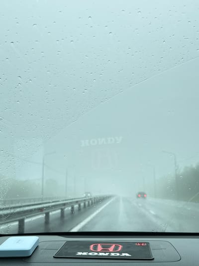 The image shows a view from inside a vehicle looking out through a rain-streaked windshield onto a highway. The weather is overcast and foggy, with visible raindrops on the glass. The road ahead is wet and appears to be a multi-lane highway with a guardrail to the left. In the distance, another vehicle with red taillights is visible. On the dashboard of the car, there is a light blue object on the left and a black mat with the Honda logo in red on the right. The Honda logo is also faintly visible on the windshield, appearing as a watermark. The overall scene suggests travel in rainy conditions on a highway. The location in the city of Пионерский, Russia, is indicated by the presence of the Honda brand, which is common in Russia, and the highway infrastructure.