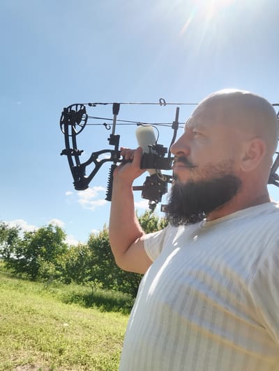 A man with a bald head and a full beard, wearing a white striped shirt, holds a black compound bow over his shoulder. He is outdoors, with a bright blue sky and the sun shining brightly, suggesting it is daytime. The background shows green trees and a grassy field, hinting at a rural or natural setting. The man is looking off to the side, with a neutral expression. The compound bow appears to be a piece of sporting equipment. The overall scene suggests an outdoor recreational activity, possibly archery practice. There is no visible text in the image.