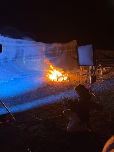 The image shows an outdoor film set at night. A large, possibly fabricated, rock-like structure is lit with blue light, appearing on the left side of the frame. Flames emanate from a metal grid or grate structure, presumably part of a special effects setup. A monitor is positioned above the flames, displaying a blue-ish image. A person stands on the right side of the frame, operating a camera and observing the scene. Cables and equipment are visible near the monitor and the camera operator, suggesting active film production. The background is dark and indistinct, with an overall low-light ambiance. San Bartolomé, Spain is not discernable from this image.