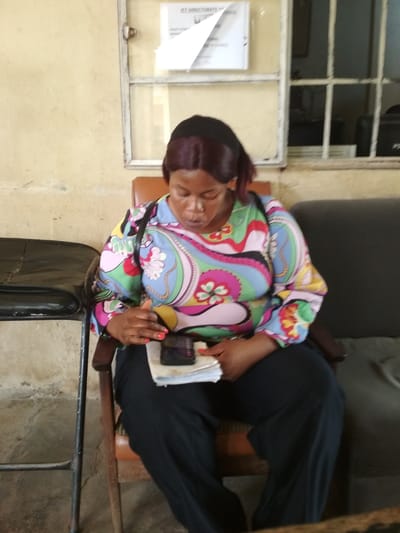 A medium shot captures a Black woman with dark hair pulled back, wearing a colorful, patterned long-sleeved blouse and black pants, seated in a wooden chair. She is looking down at a phone held in her hands, which rests on a stack of papers. The background shows a wall with a window and a bulletin board. The scene is likely indoors, possibly an office or waiting area, in Bisichi, Nigeria.
