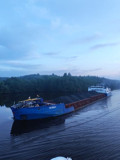 An aerial view captures a cargo barge filled with dark, granular material moving down a wide river. The barge is predominantly blue, with some orange accents on its railing and a smaller white cabin structure towards the stern. The word "БАЗАЛЬТ" is visible in white Cyrillic letters on the side of the blue hull. The river is dark and reflects the overcast sky, which is a pale blue with wispy clouds. Dense green forest lines the riverbank in the background, with a light haze obscuring the distant trees. The scene appears to be set at dawn or dusk. The location is identified as Volokov Most, Russia.