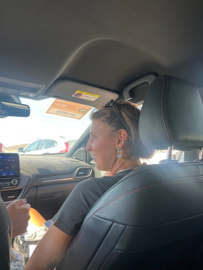 The image depicts the interior of a vehicle, specifically the front passenger compartment, during daylight hours in Parikia, Greece. The location is confirmed by a mirrored sticker on the sun visor displaying "ΕΝΟΙΚΙΑΖΟΜΕΝΑ" (Enoikiazomena), which means "for rent" in Greek, along with the year "2023."
A female individual occupies the front passenger seat, observed from behind and to her left. She has light-colored hair pulled back with sunglasses resting on her head. Her left ear is adorned with multiple piercings: a silver ear cuff on the upper helix, a small silver stud on the lower helix, and a dangling earring featuring a turquoise stone and a pale blue/white teardrop-shaped stone. She wears a dark, short-sleeved top, and a tattoo is visible on her left forearm. Her head is turned towards the left side of the vehicle, and her facial expression indicates a smile.
The vehicle's interior includes a black leather passenger seat with red stitching. The dashboard is visible, featuring a central infotainment screen. The aforementioned sun visor displays a standard yellow "AIRBAG" warning label. A partial view of another person's arm and hand is visible on the far left, suggesting the presence of a driver or another occupant. A plastic water bottle is present in the lower-left foreground. Through the windshield, a white vehicle is partially visible, indicating an outdoor setting, likely a parking area or road, under clear, bright sky conditions.