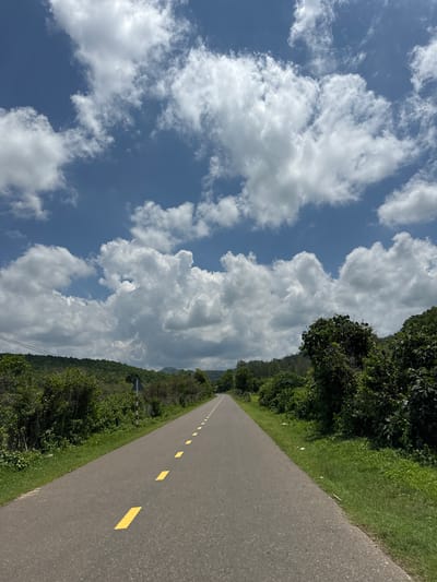The image depicts a straight, paved road with a grey asphalt surface, marked with a broken yellow line down the center. The road recedes into the distance, bordered on both sides by lush green vegetation and trees. The sky above is a vibrant blue, dotted with numerous fluffy white clouds. The scene is tranquil and rural, suggestive of a location outside of a densely populated urban area. Given the context, this scene is likely located in the city of Ham Thuan Bac, Vietnam, a region known for its natural beauty and agricultural landscapes.