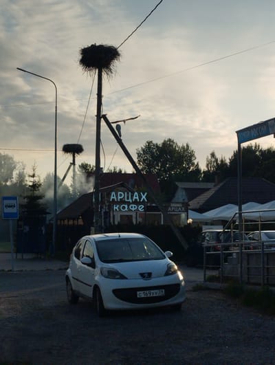 The image captures a roadside scene in Pereslavskoye, Russia, with a prominent stork's nest atop a utility pole. A white Peugeot 107 car is parked in the foreground. To the right, a blue sign reads "ПОЧТА РОССИИ" (Russian Post), and a neon sign above a building says "АРЦАХ КАФЕ" (Artsakh Cafe). Smoke is rising from a structure behind the car. The sky is overcast with clouds.