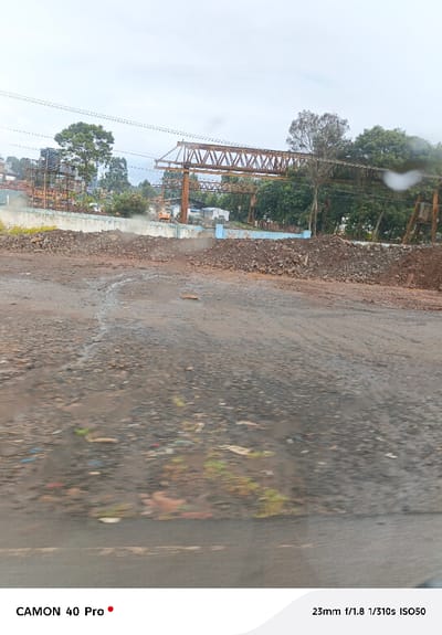 The image presents an outdoor scene depicting an industrial or construction area under an overcast sky, captured from a slightly elevated perspective, possibly from a moving vehicle given the motion blur in the foreground.
The immediate foreground is dominated by a rough, wet, and muddy ground surface, mixed with gravel and small stones. Water run-off patterns are visible across the damp earth, creating reflections and a somewhat streaky appearance. Scattered pieces of litter, including colorful plastic fragments, are embedded in the muddy surface. The lower part of the image, especially the very bottom, exhibits noticeable blur, suggesting movement during capture or a smudged/wet surface between the camera and the scene.
Moving towards the midground, large piles of reddish-brown and dark earth mixed with rocks form an uneven embankment or barrier across the frame. Behind these mounds, a low wall or fence, partially painted light blue, is visible. Beyond this, an excavator or similar yellow construction vehicle is discernible, with a faint plume of dust or exhaust rising near it. Other indistinct structures, possibly temporary buildings or storage facilities, are scattered in this area.
The background features dense green foliage from numerous trees, some with slender trunks, others forming thick clusters. Above and amidst these trees and structures, a prominent, dark brown or rusty gantry crane or a large, unfinished truss-like structure spans horizontally across the frame. This structure is characterized by its geometric, crisscrossing metal beams. Overhead utility lines or cables stretch across the sky above this gantry.
The sky is uniformly overcast and light grey, indicating a cloudy day without direct sunlight. There are also a few indistinct water droplets or smudges visible on what appears to be a windowpane in the upper right portion of the image.
The overall impression is that of a functional, possibly industrial or developing landscape, characterized by heavy machinery, earthworks, and man-made structures contrasting with natural vegetation. No specific human figures or explicit activities are clearly visible, but the presence of construction equipment implies ongoing or recent work.
At the bottom left corner of the image, the text "CAMON 40 Pro" is present. In the bottom right corner, camera metadata is displayed: "23mm f/1.8 1/310s ISO50".