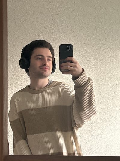 An indoor scene within the city of Hagen, Germany, depicts a young adult male reflected in a mirror. He possesses dark, wavy hair and light facial stubble. He is wearing black over-ear headphones and a ribbed knit sweater with horizontal stripes alternating between beige and light brown/taupe, layered over a grey crew-neck t-shirt. The man is holding a dark-colored smartphone with a dual-lens camera system in his right hand, oriented as if capturing his own reflection. His gaze is directed to the right of the frame, accompanied by a subtle smile. The background consists of a light-colored, textured wall. A dark wooden or plastic mirror frame is visible along the bottom and left edges of the reflection. The specific location within Hagen, Germany, is not discernible from the visual information presented.
