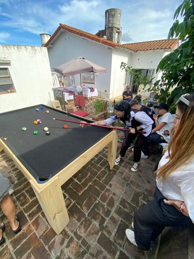A medium, angled shot captures a gathering around a pool table in what appears to be an outdoor space, potentially a patio or backyard in Monte Grande, Argentina. Several individuals surround the pool table, watching someone take a shot. The pool table is a light wood construction with a black felt surface and standard billiard balls positioned. The location features brick or stone paving. In the background, there are white-painted structures, possibly houses or buildings, with visible architectural details such as a water tower and a sunshade over outdoor seating. Laundry can be seen hanging out to dry on a line. The sky, visible in the top-left corner, is blue with some clouds. The overall atmosphere seems casual and social.